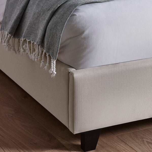 Beige upholstered bed frame with a gray blanket on a wooden floor