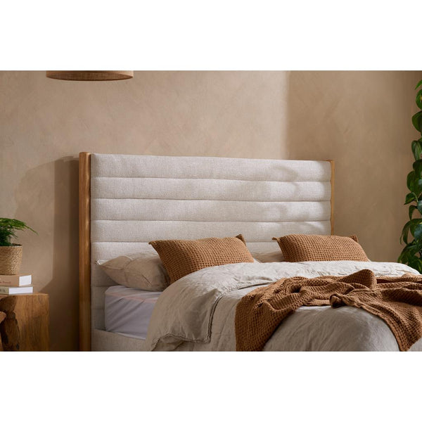 Bed with beige upholstered headboard and brown pillows against a beige wall.