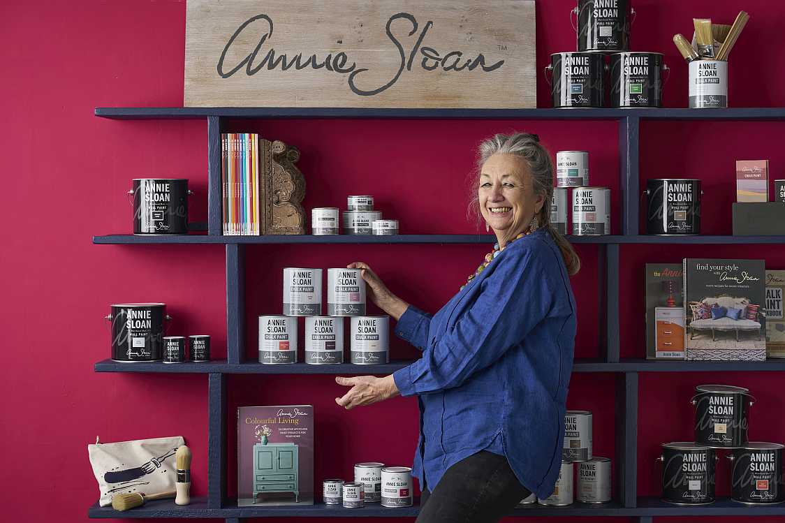 Annie Sloan standing in front of a shelf displaying Annie Sloan products against a red wall.