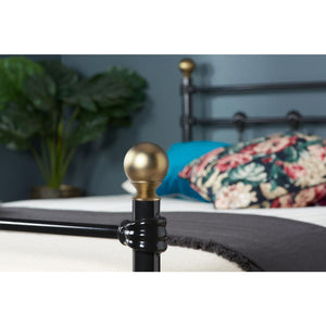 Close-up of a bedpost with gold and black design, blurred background with pillows and plant.