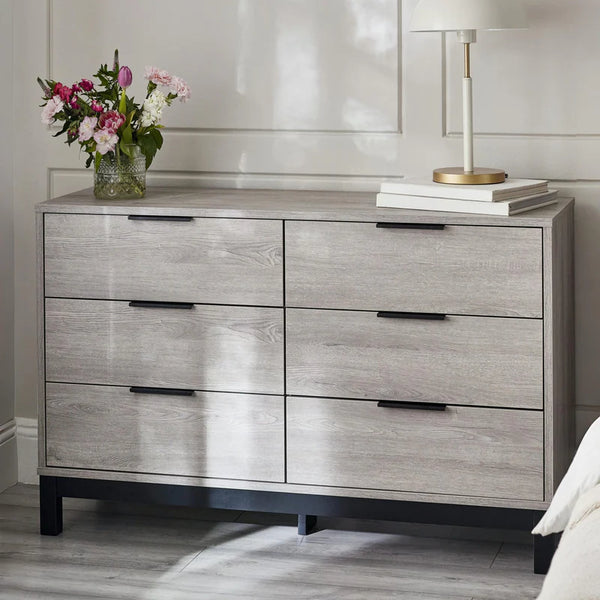 Wooden dresser with four drawers, a vase of flowers, and a lamp on top in a room setting.