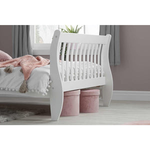 White bed frame with pink bedding and hat boxes in a room with a window and curtains.