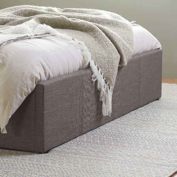 Gray upholstered bed with white bedding and a beige throw blanket on a neutral background
