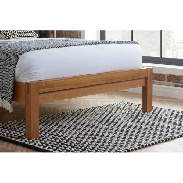 Wooden bed frame with white bedding and a patterned throw in a room with large windows.