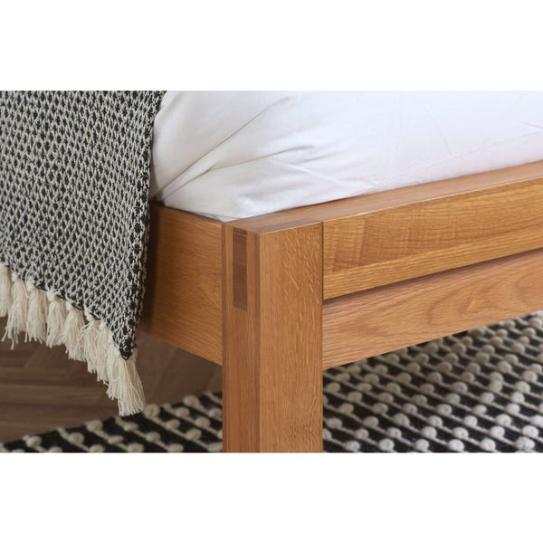 Wooden bed frame with a white mattress and black and white patterned blanket on a textured rug.