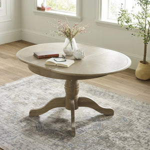 Round wooden table with books and a vase on a patterned rug in a room with windows and plants.