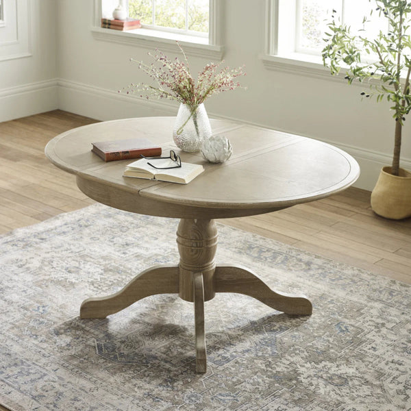Round wooden table with books and a vase on a patterned rug in a room with windows and plants.