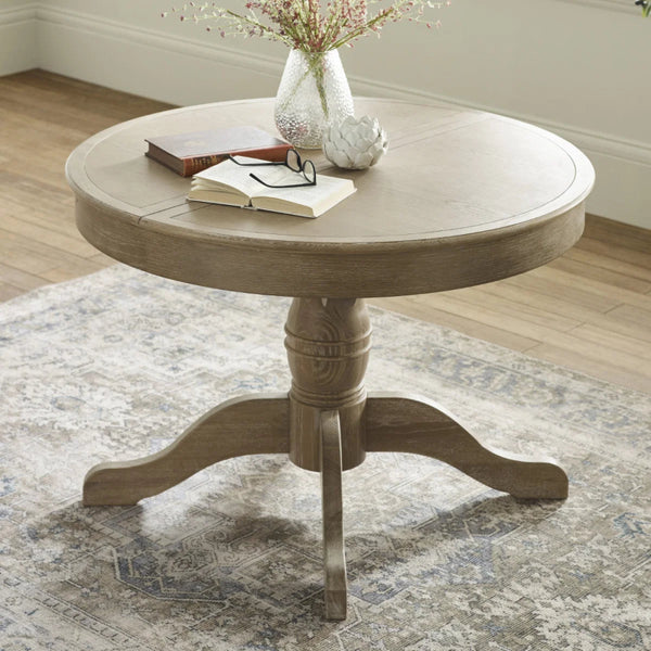 Round wooden table with a vase, book, and glasses on a patterned rug.