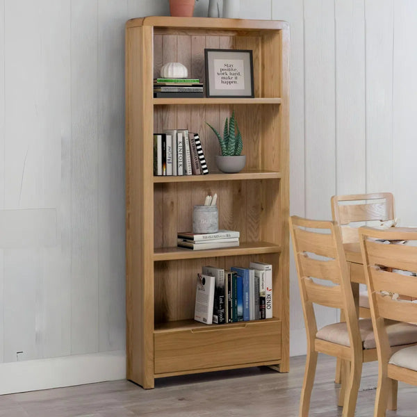 Wooden bookshelf with books and decor items against a white wall, with chairs and a table in the foreground.