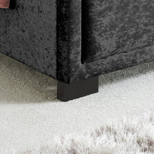 Close-up of a black velvet bed with a black foot on a carpeted floor.