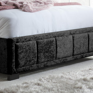 Black upholstered bed frame with white bedding and pink blanket on a carpeted floor.