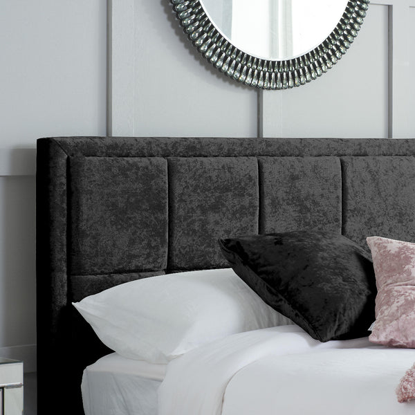 Black upholstered headboard with pillows on a bed against a white wall with a decorative mirror.