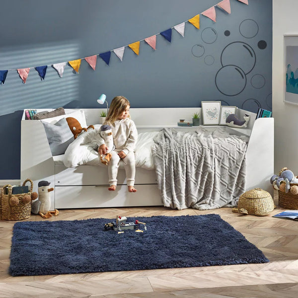 Child sitting on a bed in a bedroom with a blue rug and colorful flags on the wall.