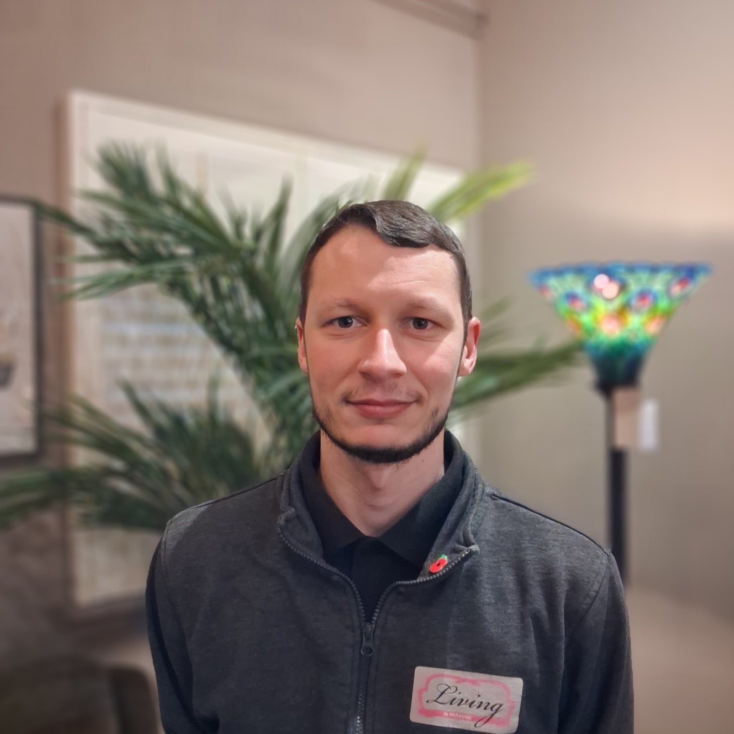 Man standing indoors with a blurred background featuring plants and a colorful lamp.