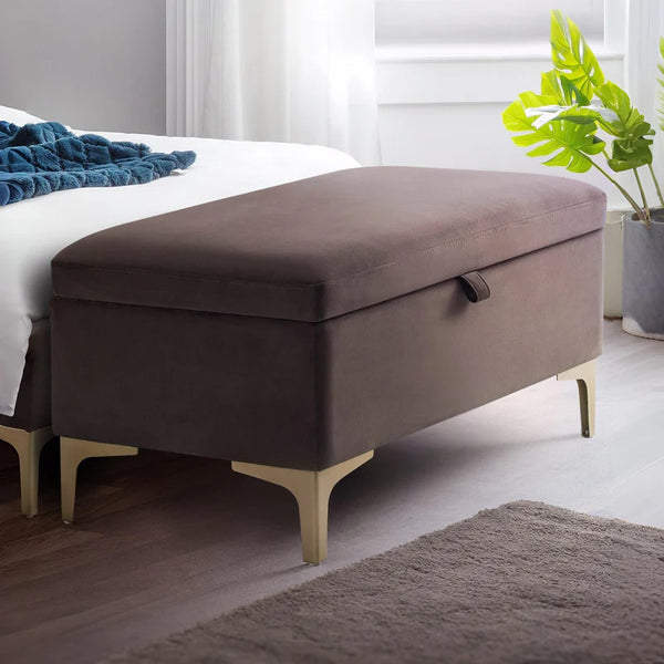 Brown storage ottoman in a bedroom setting with a bed and plant in the background.