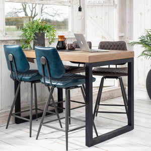 Wooden dining table with blue bar stools in a bright room with large windows.