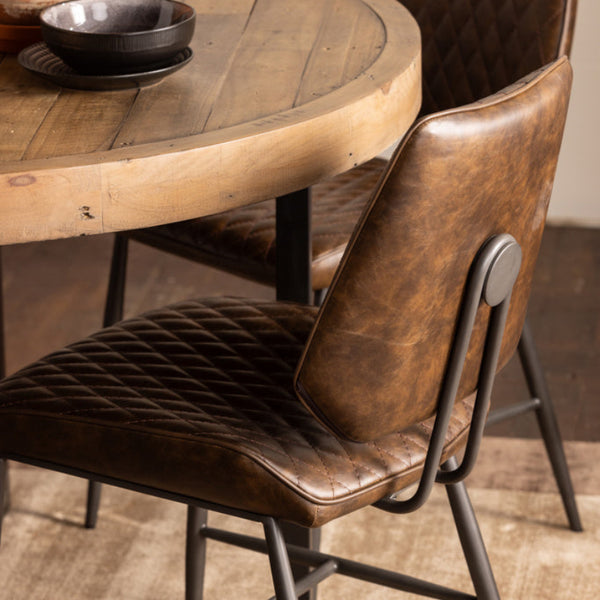 Round wooden dining table with two brown leather chairs in a room setting.