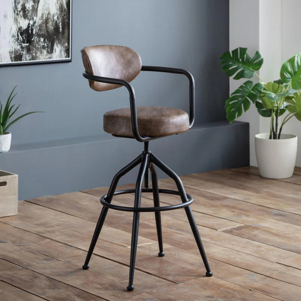 Brown bar stool with black metal frame in a room with plants and wooden floor.