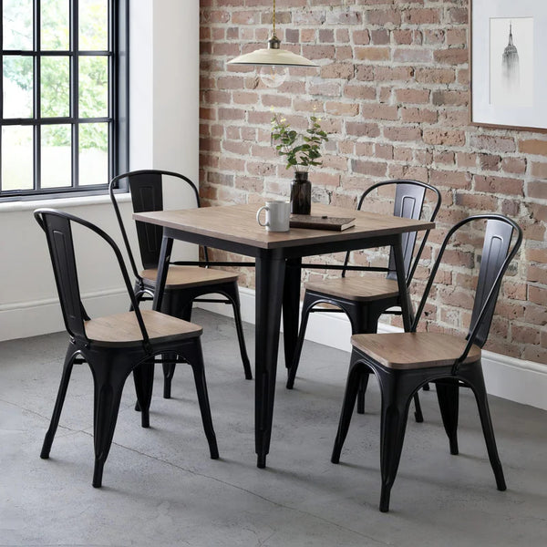 Dining area with a square table and four black metal chairs with wooden seats against a brick wall.