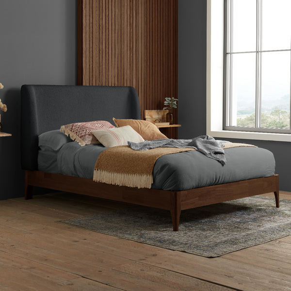 Bedroom with a bed featuring a dark upholstered headboard and wooden frame, with pillows and blankets on a wooden floor.