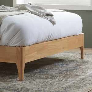 Wooden bed frame with white bedding and gray blanket on a patterned rug.