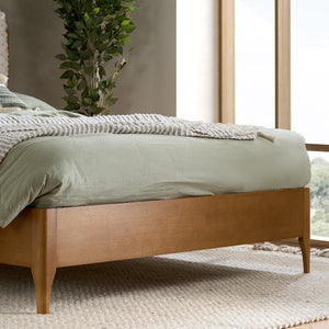 Wooden bed with green bedding in a room with large windows and a plant.