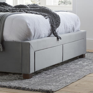 Gray upholstered bed with storage drawers in a room with a rug and window.