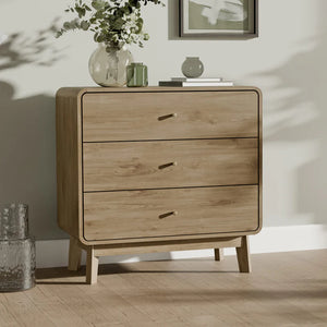 Wooden dresser with three drawers on a wooden floor, with a vase of flowers and a framed picture in the background.