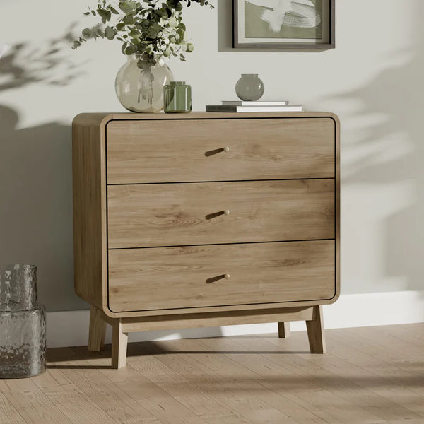 Wooden dresser with three drawers on a wooden floor, with a vase of flowers and a framed picture in the background.