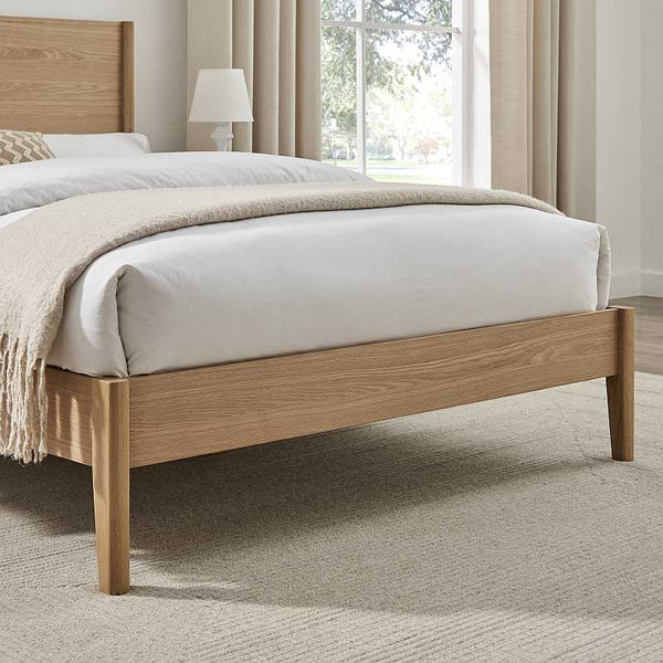 Wooden bed with white bedding and beige blanket in a room with curtains and a lamp.