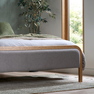 Gray upholstered bed with wooden frame in a room with a plant and window.