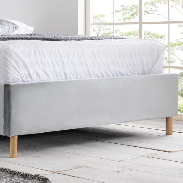 Gray upholstered bed with wooden legs in a bright room with large windows.