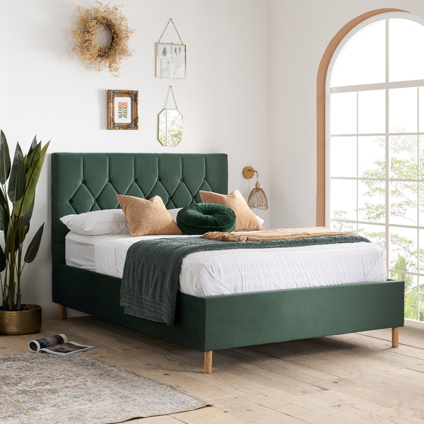 Green upholstered bed in a bright room with large windows and decorative elements.