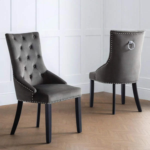 Two gray upholstered chairs with nailhead trim and a kncoker on the back of the chair on a wooden floor.
