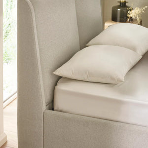 Beige upholstered bed with pillows and a lamp on a nightstand in a bright room.