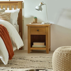 Wooden bedside table with lamp and books next to a bed with white bedding and orange throw.
