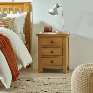 Wooden nightstand with three drawers next to a bed with white bedding and an orange throw blanket.