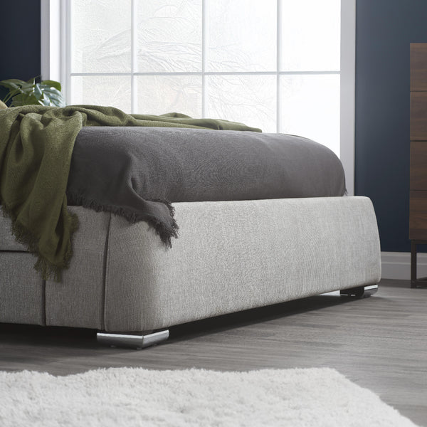 Gray upholstered bed with a green blanket in a room with large windows.