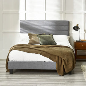 Gray upholstered bed with brown blanket and pillows against a white paneled wall.