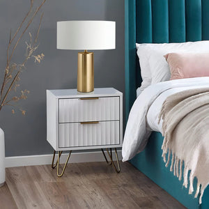 White nightstand with gold legs and lamp next to a bed with teal headboard and white bedding.