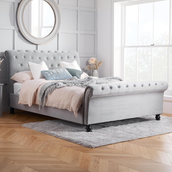 Gray upholstered bed in a bedroom with wooden flooring and a large window.