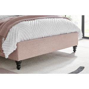 Beige upholstered bed with white textured blanket and pink throw in a room with large windows.