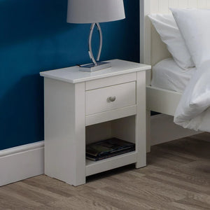 White nightstand with a lamp and books next to a bed with white bedding.