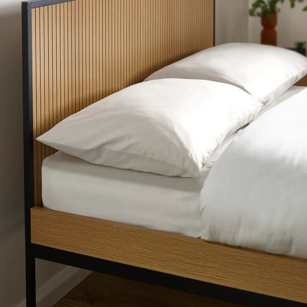 Wooden bed frame with white bedding and pillows against a neutral wall.