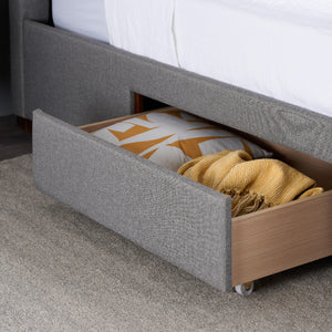 Gray upholstered bed with open drawer containing folded yellow blanket and patterned pillow.