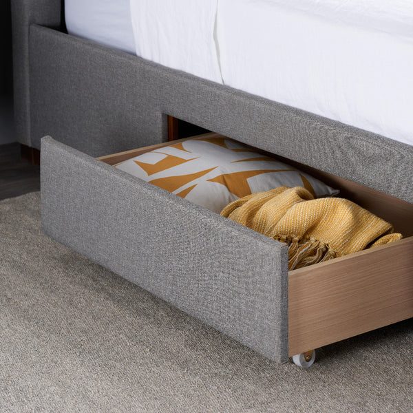 Gray upholstered bed with open drawer containing folded yellow blanket and patterned pillow.