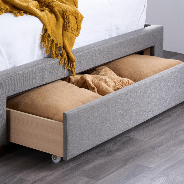 Gray upholstered bed with open drawer containing pillows and blankets on a wooden floor.