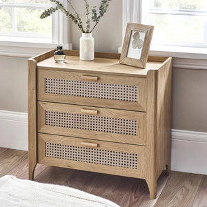 Wooden chest of drawers with decorative patterns on drawers, placed in a room with a window and plant.