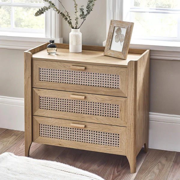 Wooden chest of drawers with decorative patterns on drawers, placed in a room with a window and plant.