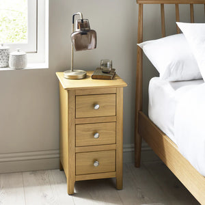 Wooden bedside table with lamp and drawers next to a bed in a bedroom.